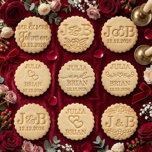 May include: A collection of round, scalloped-edge cookies with various embossed designs and text. The cookies feature names, initials, and dates, including "MR & MRS Johnson 12.12.2026" and "J & B 12.12.2026". The cookies are arranged on a red surface with floral accents.