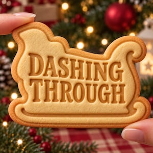 May include: A light tan, sleigh-shaped cookie featuring the embossed text DASHING THROUGH. The cookie has a smooth, baked surface with defined edges and a raised border, showcasing a clear, legible font centered within the sleigh silhouette.