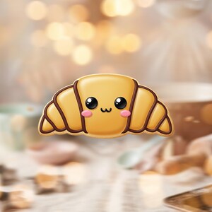 May include: A cartoon-style croissant with a smiling face, black eyes, and pink cheeks. The pastry is golden brown with darker brown accents, giving it a baked appearance. The background is blurred with warm, soft lighting.