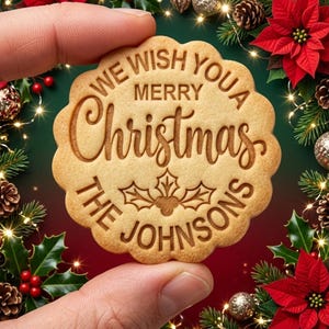 May include: A round, scalloped-edge cookie with a light brown surface featuring the embossed text "WE WISH YOU A MERRY CHRISTMAS" and "THE JOHNSONS" surrounding a central holly leaf and berry illustration.