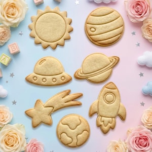 May include: A collection of seven space-themed sugar cookies shaped like a sun, a planet with rings, a rocket ship, a shooting star, a UFO, a cratered planet, and a striped planet, arranged on a gradient blue and pink background with roses.