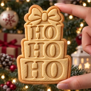 May include: A light tan, rectangular-shaped cookie with rounded edges, featuring a molded design of three stacked gift boxes topped with a bow. The text HO HO HO is embossed in a bold, serif font across the three gift box sections.