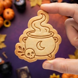 May include: A light tan cookie shaped like a cauldron with a crescent moon emblem on the front, steam rising from the top, and a small skull and leaf detail at the base. The cookie has a smooth, baked texture with embossed lines defining the design.