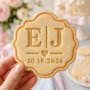 May include: A round, scalloped-edge sugar cookie featuring an embossed design with the letters E and J separated by a vertical line, a small heart icon below, and the date 10.18.2026 printed at the bottom in a clear, sans-serif font.