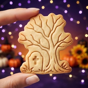 May include: A light tan, flat, die-cut cookie shaped like a tree with a tombstone at its base. The cookie features embossed details of a bare-branched tree and a tombstone with a cross symbol centered on the front surface of the baked dough.
