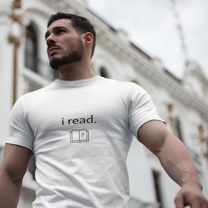 Puede incluir: Camiseta blanca con el texto "i read." y un pequeño dibujo de un libro abierto. La camiseta es de manga corta y parece ser de algodón. El fondo es una escena exterior borrosa con un edificio.
