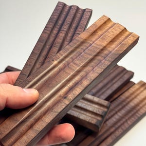 May include: A collection of rectangular wooden soap dishes, each featuring a grooved design. The dishes are crafted from dark brown wood, showcasing a natural wood grain. The grooves are parallel and span the width of the dish.