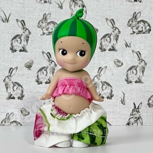 May include: A whimsical figurine of a baby-like character dressed as a watermelon. The figure wears a green and pink watermelon-themed outfit, including a green striped cap. The background features a repeating pattern of rabbits.