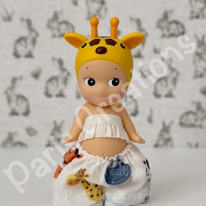 May include: A small doll wearing a yellow giraffe hat and a white ruffled top and pants. The pants are decorated with giraffe and blue pouch designs. The doll has a light skin tone and is set against a background with a rabbit pattern.