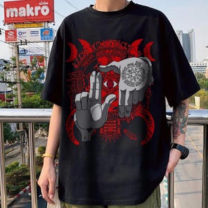 May include: Black t-shirt featuring a red and gray graphic design. The design includes two hands framing a red eye, surrounded by a circular pattern with text. The word "HELLSING" is visible in the design.
