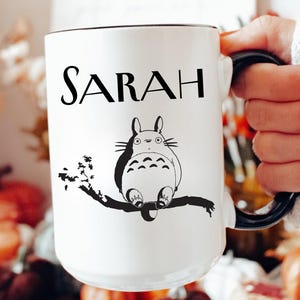 May include: White ceramic mug with the name "SARAH" in black lettering. A black and white illustration of a cartoon character is below the name. The mug has a black handle and rim.