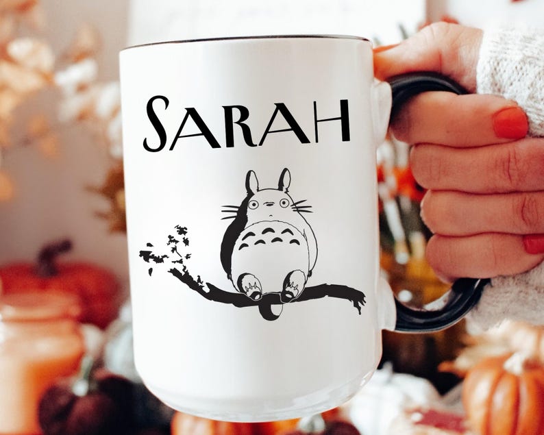 May include: White ceramic mug with the name "SARAH" in black lettering. A black and white illustration of a cartoon character is below the name. The mug has a black handle and rim.