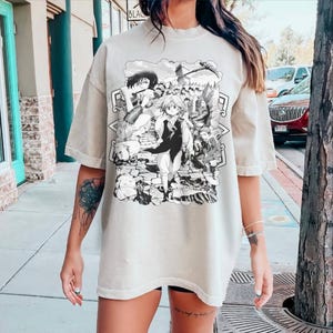 May include: Oversized beige t-shirt featuring a black and white anime graphic. The graphic depicts multiple characters in a detailed, action-oriented scene. The shirt has short sleeves and a relaxed fit, suitable for casual wear.