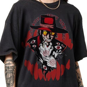 May include: Black graphic t-shirt featuring a stylized illustration of a character with a top hat, yellow glasses, and a red coat. The design includes red accents, multiple eyes, and text in a circular pattern. The shirt is a dark color.
