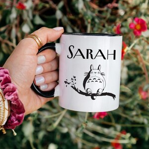 May include: White ceramic mug with a black interior and handle, featuring the name "SARAH" in black lettering. A black and white illustration of a cartoon character is also on the mug. The mug is held by a person.