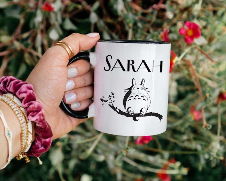 May include: White ceramic mug with a black interior and handle, featuring the name "SARAH" in black lettering. A black and white illustration of a cartoon character is also on the mug. The mug is held by a person.