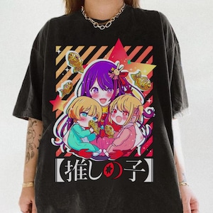 May include: Black graphic t-shirt featuring anime-style characters with purple, blonde, and blue hair. The design includes fish-shaped pastries and Japanese text. The artwork is set against a backdrop of red, yellow, and orange geometric shapes.