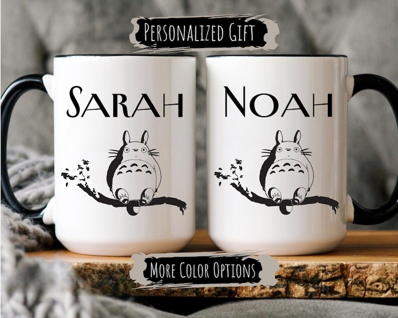 May include: Two white ceramic mugs with black handles and rims. Each mug features a black and white illustration of a cartoon character and a name. The text "SARAH" and "NOAH" are printed on the mugs. The text "PERSONALIZED GIFT" and "MORE COLOR OPTIONS" are also visible.