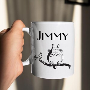 May include: White ceramic mug with the name "JIMMY" in black lettering. Below the name is a black and white illustration of a cartoon character sitting on a branch. The mug has a curved handle.