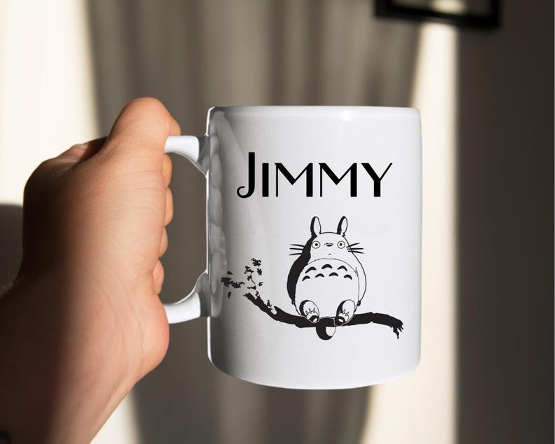 May include: White ceramic mug with the name "JIMMY" in black lettering. Below the name is a black and white illustration of a cartoon character sitting on a branch. The mug has a curved handle.