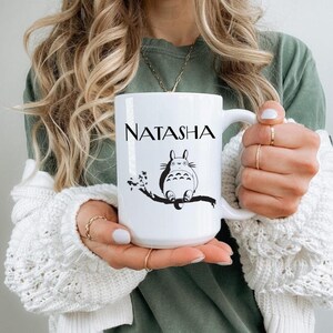May include: White ceramic mug featuring the name "NATASHA" in black lettering. Below the name is a black and white illustration of a cartoon character sitting on a tree branch. The mug has a large handle.
