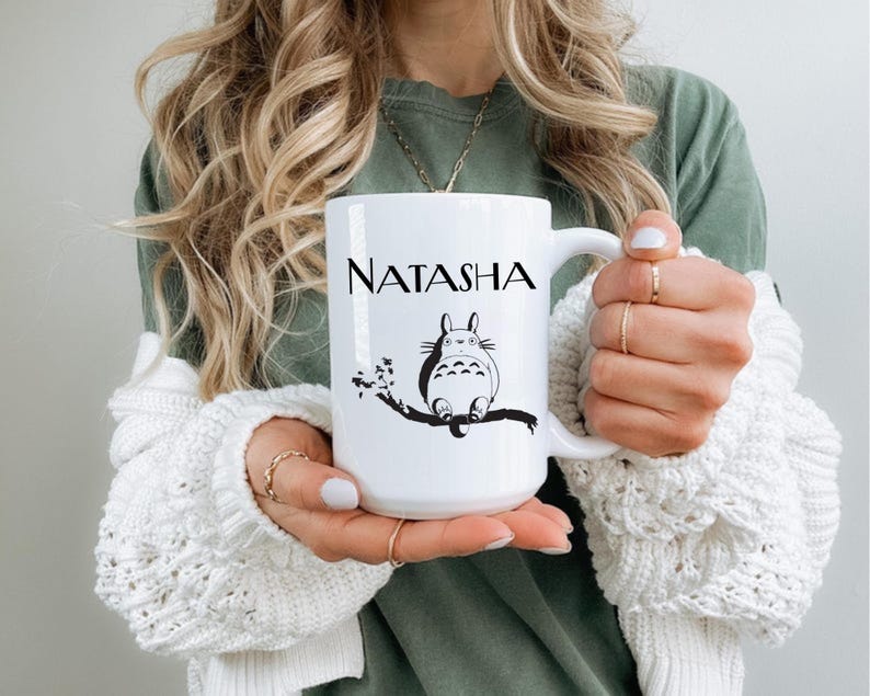 May include: White ceramic mug featuring the name "NATASHA" in black lettering. Below the name is a black and white illustration of a cartoon character sitting on a tree branch. The mug has a large handle.