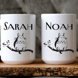 May include: Two white ceramic mugs with black text and illustrations. One mug reads "SARAH" above a cartoon character sitting on a branch. The other mug reads "NOAH" above a similar illustration. Both mugs have a handle.