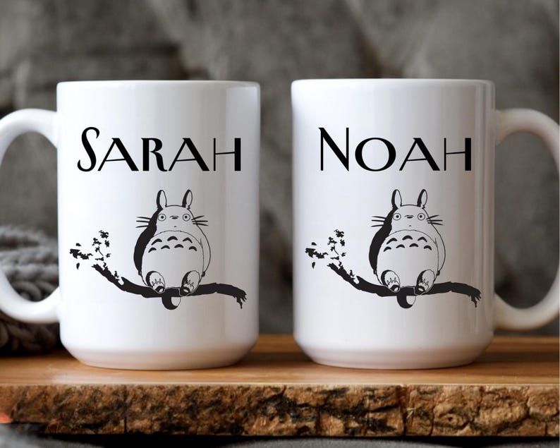 May include: Two white ceramic mugs with black text and illustrations. One mug reads "SARAH" above a cartoon character sitting on a branch. The other mug reads "NOAH" above a similar illustration. Both mugs have a handle.