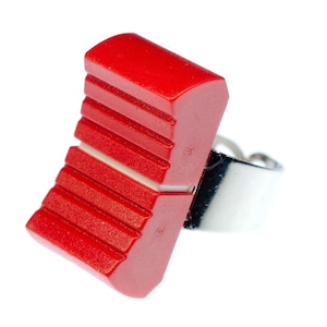 Puede incluir: Anillo rojo llamativo con un diseño rectangular texturizado. El anillo presenta una banda plateada y una forma geométrica audaz. La sección roja tiene ranuras horizontales, lo que agrega interés visual a la joya.