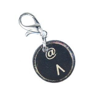 May include: A circular charm with a black face and white symbols, including an at symbol and a caret. It is attached to a silver-coloured lobster clasp. The charm is likely a pendant or decorative element for jewellery or accessories.
