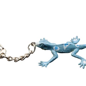May include: A light blue lizard keychain featuring a textured, scaly surface with white and dark blue circular and triangular patterns along its back. The lizard is attached to a silver-toned metal split ring by a short, linked metal chain.