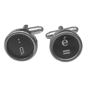 May include: Two cufflinks with a typewriter key design. Each cufflink has a black circular face with a silver metal rim and post. One shows the Greek letter beta, the other the letter 'e' with an accent and two horizontal lines.