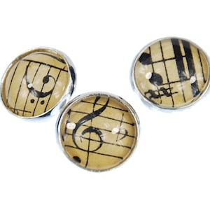 Può includere: Tre bottoni rotondi argentati con una parte superiore in vetro trasparente. Ogni bottone presenta un'immagine in bianco e nero di note musicali e spartiti su uno sfondo beige.