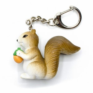 Puede incluir: Un llavero de ardilla marrón y blanco que sostiene una pequeña bellota naranja. La ardilla está hecha de plástico y tiene un diseño detallado y realista. El llavero está unido a un anillo de metal plateado.