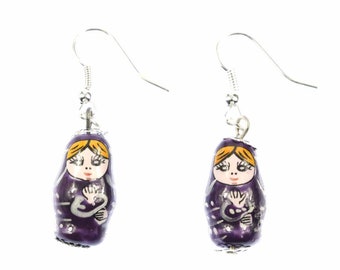 May include: Purple and white painted wooden earrings shaped like Russian nesting dolls with yellow hair and painted faces. The earrings have silver hooks.
