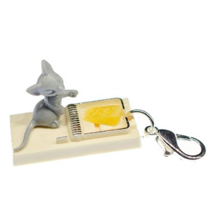 Könnte beinhalten: Eine kleine graue Mausfigur, die in einer Miniatur-Mausefalle mit einem gelben Stück Käse gefangen ist. Die Mausefalle ist an einem silbernen Schlüsselanhänger befestigt.