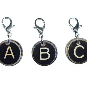 May include: Three circular charms displaying the letters A, B, and C. Each charm features a black backdrop with a white letter and a silver-coloured clasp. These charms are likely for a bracelet or necklace.
