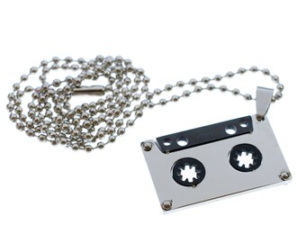 May include: A silver chain necklace with a silver and black cassette tape pendant. The pendant has a black plastic insert and two silver circles.