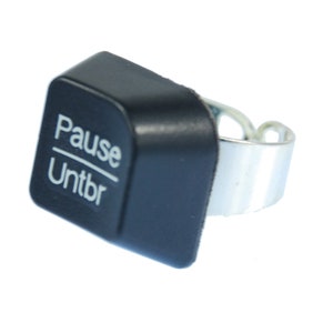May include: Black plastic ring with a silver metal band. The ring has a square button with the words "Pause" and "Untbr" printed on it.