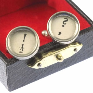 Puede incluir: Una caja de gemelos de terciopelo negro y rojo con dos gemelos de plata. Un gemelo tiene un signo de interrogación y un punto y coma, el otro tiene un signo de exclamación y la fracción 1/2.