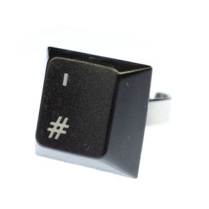 Puede incluir: Un anillo de plástico negro con una banda plateada. El anillo tiene la forma de una tecla de computadora con los símbolos "#" y "|" impresos en él.
