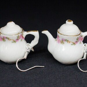Puede incluir: Un par de pendientes de porcelana blanca en forma de tetera con diseños florales rosas y detalles dorados. Los pendientes tienen ganchos plateados.