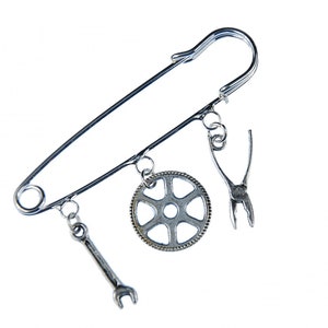 May include: A silver safety pin with three charms: a wrench, a gear, and a pair of pliers.