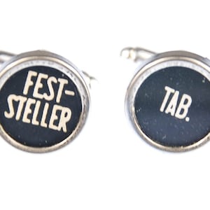 May include: A pair of silver cufflinks with black enamel. One cufflink has the text "FEST-STELLER" in white lettering. The other cufflink has the text "TAB." in white lettering.