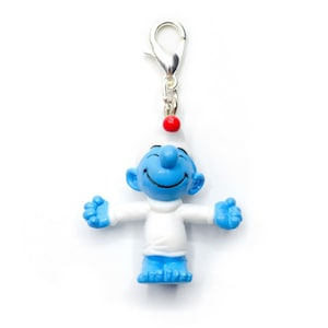 Könnte beinhalten: Eine blaue Schlumpf-Figur mit einem weißen Hut und einem roten Bommel. Der Schlumpf lächelt und hat die Arme ausgestreckt. Die Figur ist an einem silbernen Anhänger befestigt.