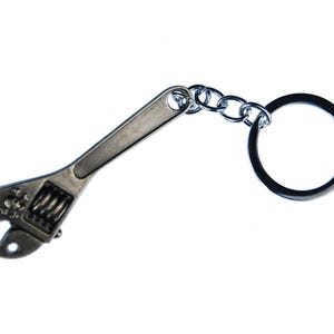Puede incluir: Un llavero de llave inglesa de color plateado. La llave inglesa en miniatura está unida a una cadena y a un llavero negro. La llave inglesa tiene un diseño de calavera en la cabeza.