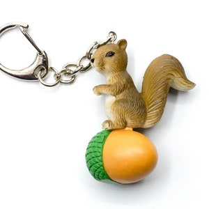 Puede incluir: Un llavero de ardilla marrón con cola tupida, posada sobre una bellota verde y naranja. El llavero tiene un anillo de metal plateado.