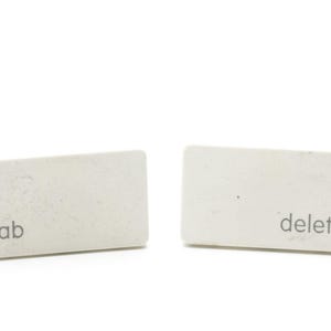 May include: Two rectangular white keyboard keys positioned side by side. The left key features the lowercase text tab printed in a dark gray sans-serif font, while the right key features the lowercase text delete in the same dark gray font.