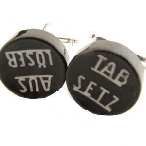 May include: A pair of black cufflinks with silver-tone fittings. Each circular cufflink features white text. One reads "AUS LOSER" and the other "TAB SETZ".