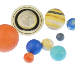Op de afbeelding: Een verzameling van negen bolvormige objecten van verschillende groottes en kleuren, waaronder een grote oranje bol, een crèmekleurige bol met een ring, een geel en bruin gestreepte bol, een blauwe bol, een kleine grijze bol en diverse kleinere bollen.
